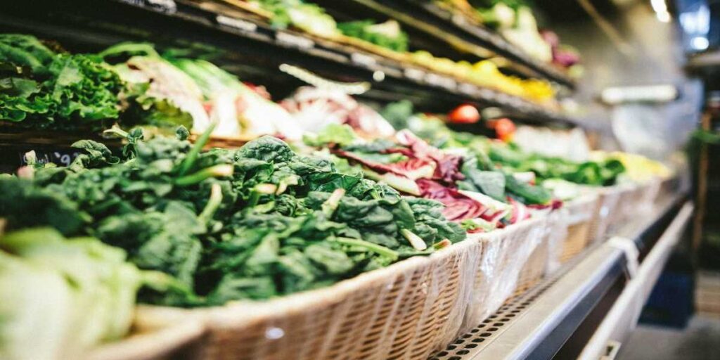 A basket of vegetables in the foreground with other produce behind it.